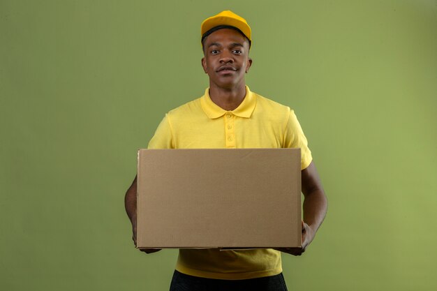 Entrega hombre afroamericano en polo amarillo y gorra de pie con caja de cartón grande mirando confiado en verde