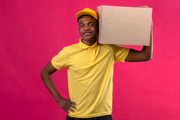 Entrega hombre afroamericano en polo amarillo y gorra de pie con caja de cartón en el hombro sonriendo amable en rosa