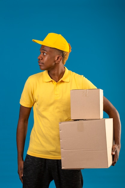Entrega hombre afroamericano en polo amarillo y gorra de pie con cajas de cartón mirando a un lado con cara seria en azul aislado