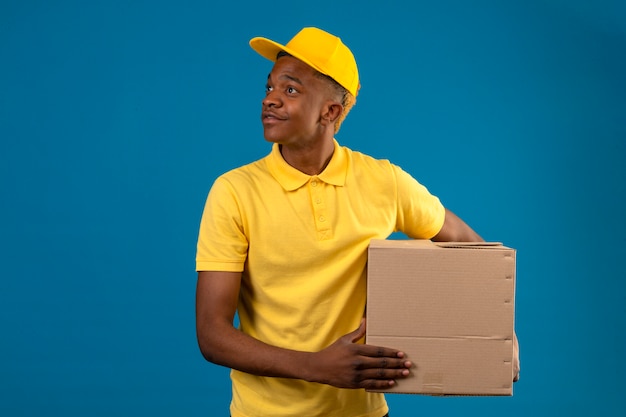 Entrega hombre afroamericano en polo amarillo y gorra de pie con cajas de cartón mirando a un lado con una sonrisa en la cara en azul aislado