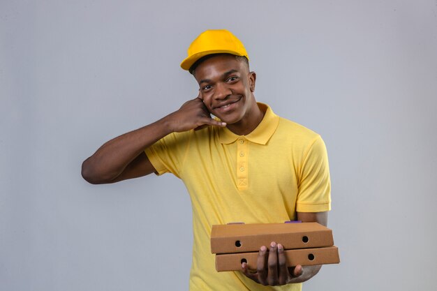 Entrega hombre afroamericano en polo amarillo y gorra de pie con cajas de pizza haciendo gesto de llamarme mirando confiado sonriendo amable