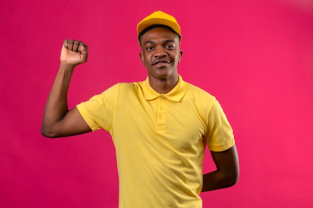 Entrega hombre afroamericano en polo amarillo y gorra que parece seguro con una sonrisa en la cara levantando el puño después de un concepto de ganador de la victoria en rosa aislado