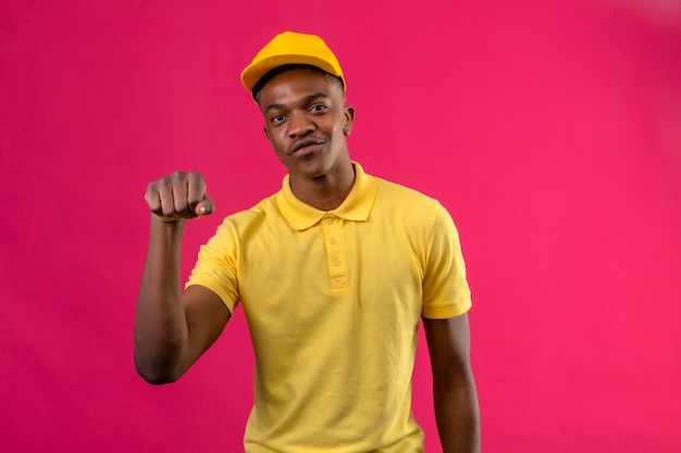 Entrega hombre afroamericano en polo amarillo y gorra que parece seguro con una sonrisa en la cara levantando el puño en rosa aislado