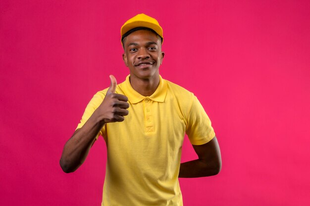 Entrega hombre afroamericano en polo amarillo y gorra sonriendo amigable mostrando los pulgares para arriba en rosa