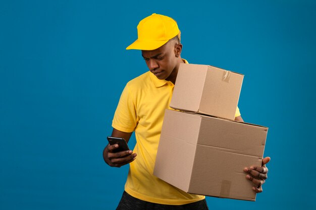 Entrega hombre afroamericano en polo amarillo y gorra sosteniendo cajas de cartón mirando la pantalla de su teléfono móvil de pie en azul