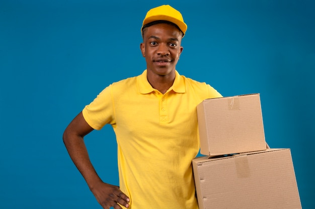 Entrega hombre afroamericano en polo amarillo y gorra sosteniendo cajas de cartón con sonrisa amistosa de pie en azul aislado