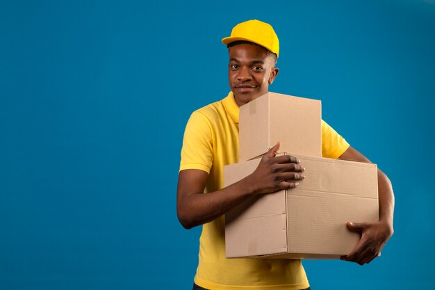 Entrega hombre afroamericano en polo amarillo y gorra sosteniendo cajas de cartón con sonrisa amistosa de pie en azul aislado