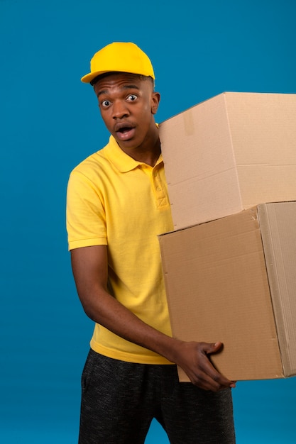 Entrega hombre afroamericano en polo amarillo y gorra sosteniendo cajas de cartón sorprendido con cara de sorpresa de pie en azul aislado