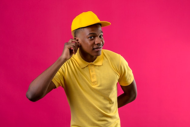 Entrega hombre afroamericano en polo amarillo y gorra sosteniendo la mano cerca de su oreja tratando de escuchar la conversación de alguien de pie en rosa