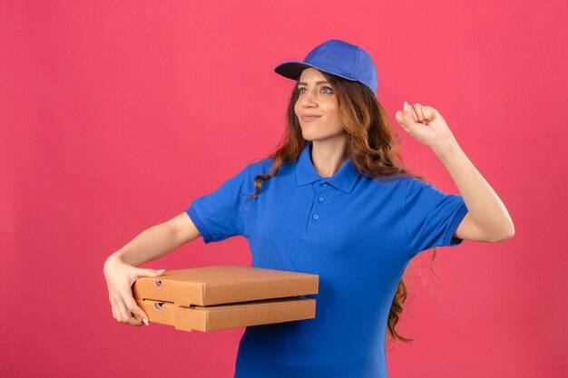 Entrega joven mujer con cabello rizado con camisa polo azul y gorra de pie con cajas de pizza levantando el puño después de una victoria concepto de ganador de cara feliz sobre fondo rosa aislado
