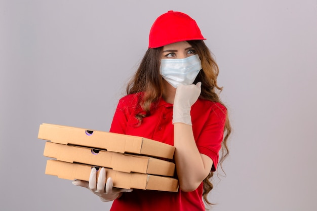 Entrega joven mujer con cabello rizado con camisa polo roja y gorra en máscara protectora médica y guantes de pie con cajas de pizza pensando concentrado en la duda con la mano en la barbilla sobre aislado