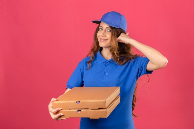 Entrega joven mujer con cabello rizado vistiendo polo azul y gorra de pie con cajas de pizza haciendo gesto de llamarme mirando confiado sobre fondo rosa aislado