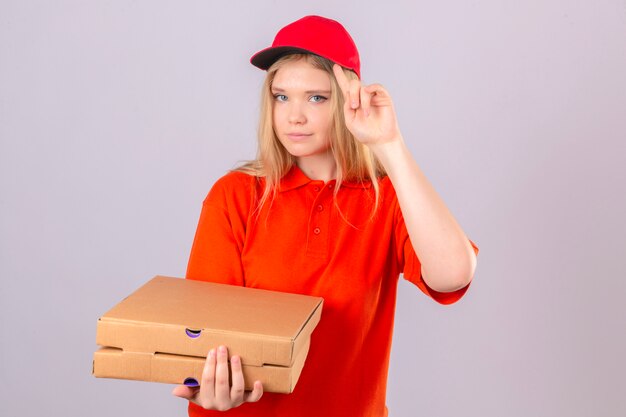 Entrega joven mujer en camisa polo naranja y gorra roja apuntando su gorra con el dedo pensando centrado en una tarea sobre fondo blanco aislado
