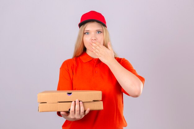 Entrega joven mujer en camisa polo naranja y gorra roja sosteniendo cajas de pizza mirando sorprendido cubriendo la boca con la mano sobre fondo blanco aislado