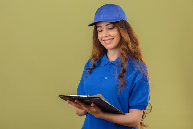 Entrega joven mujer con pelo rizado vistiendo polo azul y gorra mirando el portapapeles en manos sonriendo con cara feliz sobre fondo verde aislado