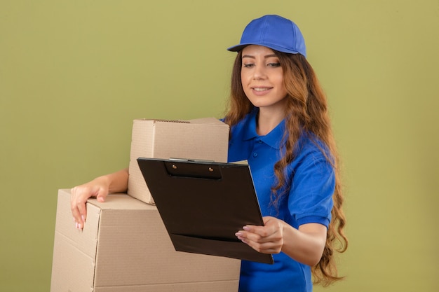 Entrega joven mujer con pelo rizado vistiendo polo azul y gorra de pie con cajas de cartón mirando el portapapeles sonriendo sobre fondo verde aislado