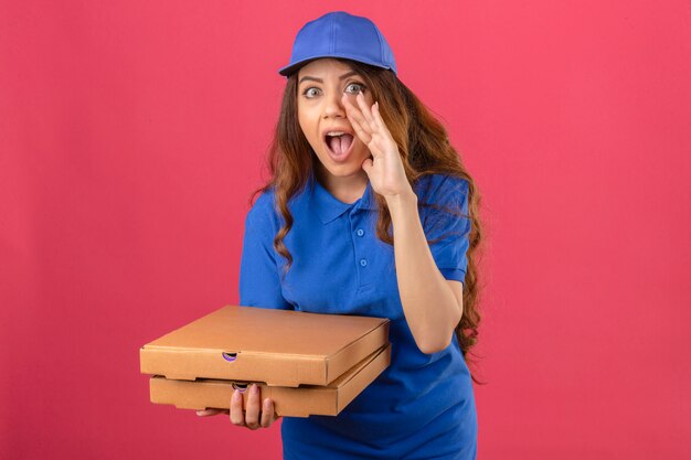 Entrega joven mujer con pelo rizado vistiendo polo azul y gorra de pie con cajas de pizza gritando con la boca abierta sobre fondo rosa aislado