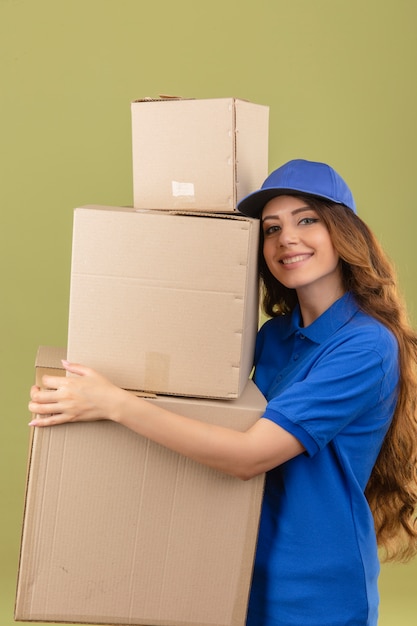 Foto gratuita entrega joven mujer con pelo rizado vistiendo polo azul y gorra de pie con pila de cajas de cartón en manos sonriendo amable sobre fondo verde aislado