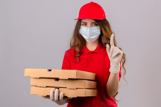 Entrega joven mujer con pelo rizado vistiendo polo rojo y gorra en máscara protectora médica y guantes de pie con cajas de pizza apuntando con el dedo hacia arriba con cara seria sobre backgrou blanco aislado