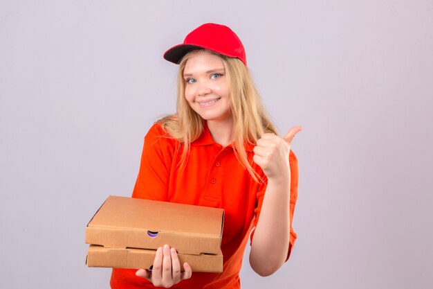 Entrega joven mujer en polo naranja y gorra roja de pie con cajas de pizza mostrando el pulgar hacia arriba sonriendo alegremente sobre fondo blanco aislado