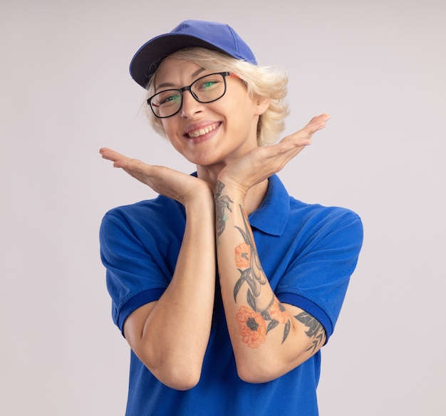 Entrega joven mujer en uniforme azul y gorra feliz y positiva sonriendo alegremente de pie sobre la pared blanca