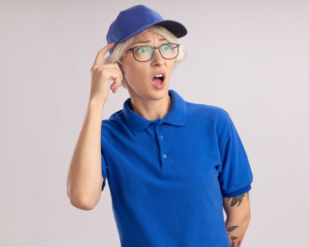 Entrega joven mujer en uniforme azul y gorra con gafas mirando a un lado sorprendido y preocupado de pie sobre la pared blanca