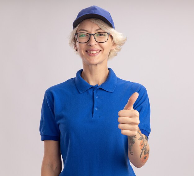 Entrega joven mujer en uniforme azul y gorra con gafas mirando sonriendo alegremente mostrando los pulgares para arriba de pie sobre la pared blanca