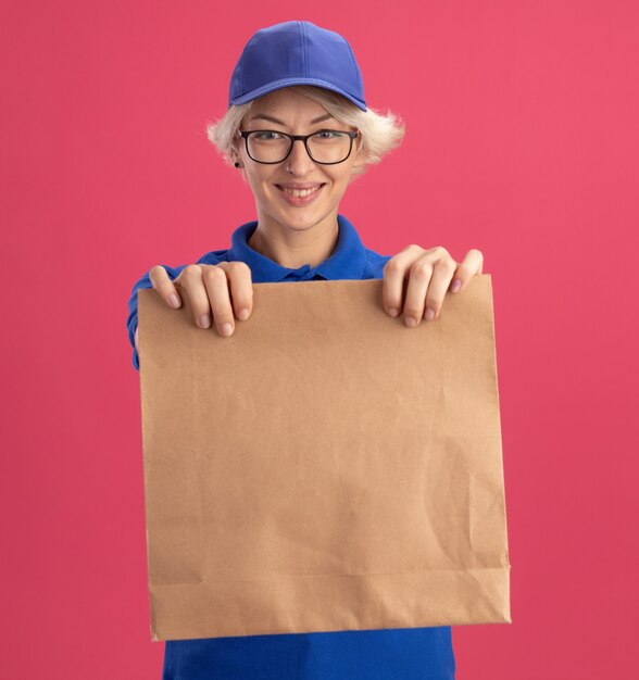 Entrega joven mujer en uniforme azul y gorra con gafas sosteniendo el paquete de papel mirando sonriendo alegremente sobre la pared rosa