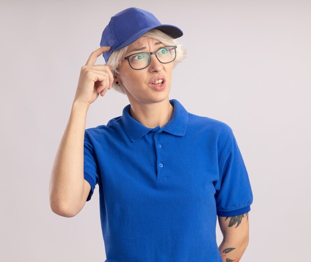 Entrega joven mujer en uniforme azul y gorra mirando a un lado desconcertado rascarse la cabeza con el dedo parado sobre la pared blanca