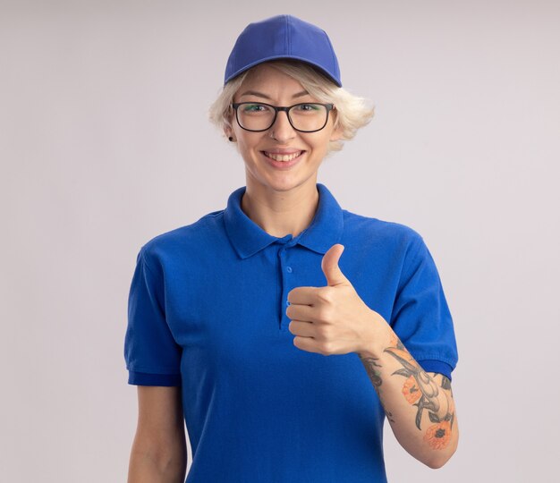 Entrega joven mujer en uniforme azul y gorra mirando sonriendo alegremente mostrando los pulgares para arriba de pie sobre la pared blanca