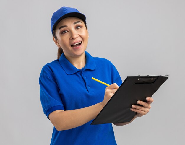 Entrega joven mujer en uniforme azul y gorra sosteniendo portapapeles y bolígrafo escribiendo algo con una sonrisa en la cara de pie sobre la pared blanca