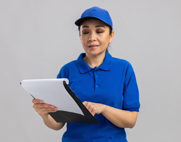 Entrega joven mujer en uniforme azul y gorra sosteniendo el portapapeles mirándolo sonriendo confiado de pie sobre la pared blanca