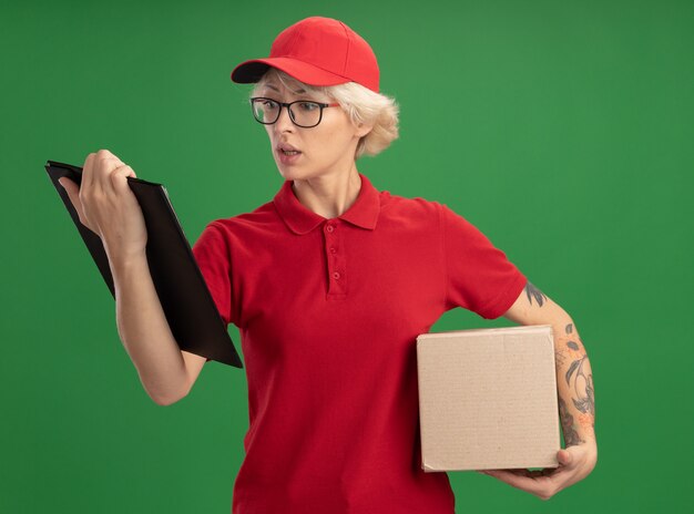 Entrega joven mujer en uniforme rojo y gorra con gafas con caja de cartón mirando el portapapeles preocupado de pie sobre la pared verde