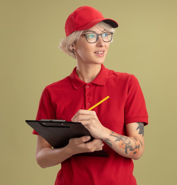 Entrega joven mujer en uniforme rojo y gorra con gafas con portapapeles y lápiz mirando a un lado con una sonrisa en la cara de pie sobre la pared verde