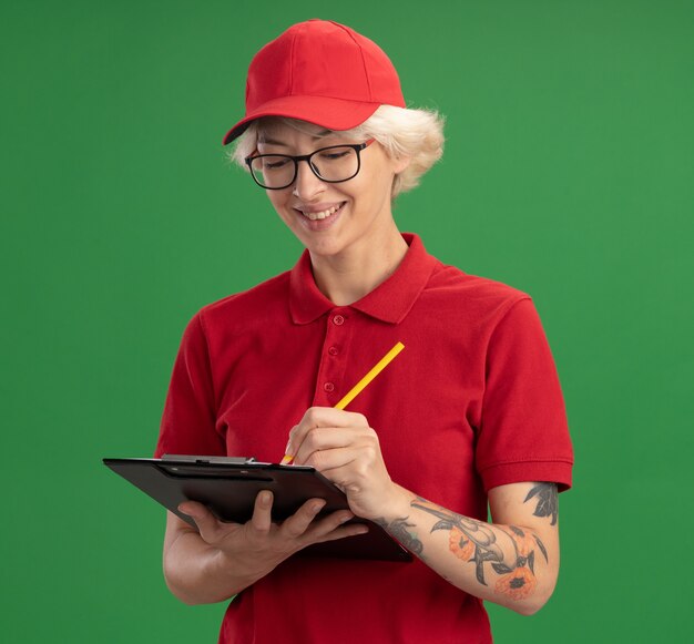 Entrega joven mujer en uniforme rojo y gorra con gafas con portapapeles con páginas en blanco y lápiz escribiendo algo con una sonrisa en la cara de pie sobre la pared verde