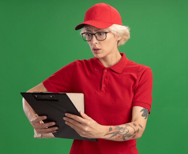 Entrega joven mujer en uniforme rojo y gorra con gafas sosteniendo una caja de cartón con portapapeles mirándolo preocupado de pie sobre la pared verde