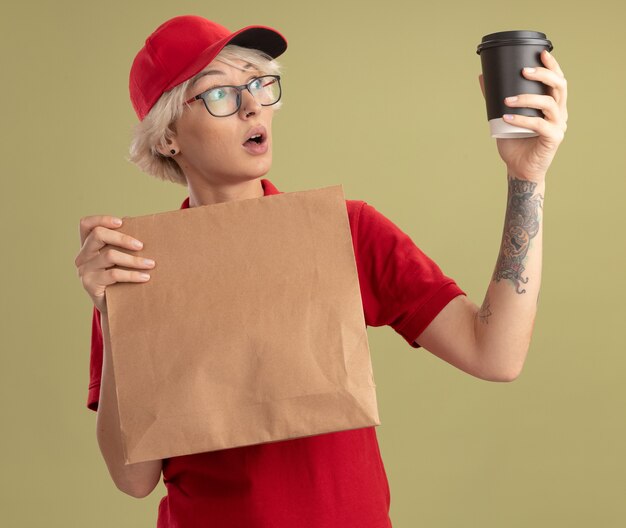 Entrega joven mujer en uniforme rojo y gorra con gafas sosteniendo el paquete de papel mirando la taza de café en su mano lookign sorprendido y asombrado de pie sobre la pared verde