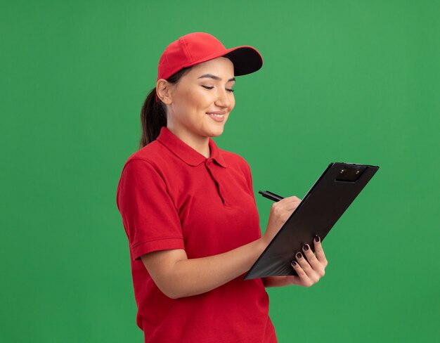 Entrega joven mujer en uniforme rojo y gorra con portapapeles y lápiz escribiendo sonriendo alegremente de pie sobre la pared verde