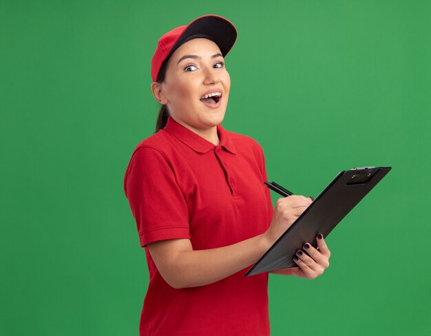 Entrega joven mujer en uniforme rojo y gorra con portapapeles y lápiz mirando al frente feliz y emocionado de pie sobre la pared verde