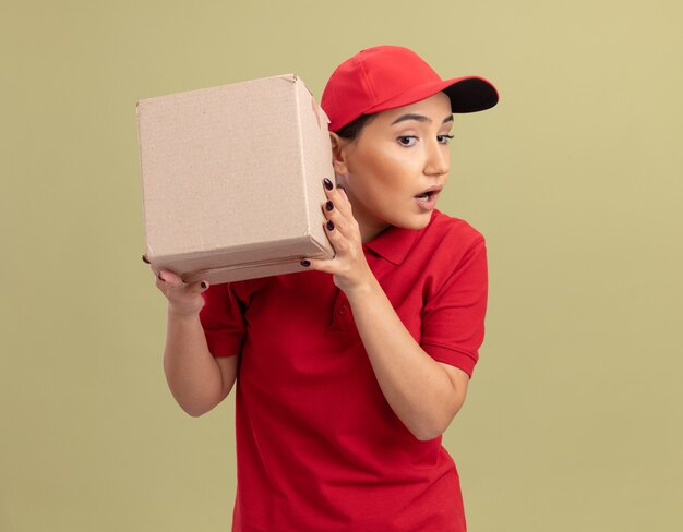 Entrega joven mujer en uniforme rojo y gorra sosteniendo una caja de cartón sobre su oreja escuchando parado sobre pared verde