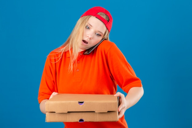 Entrega joven mujer vestida con camisa polo naranja y gorra roja sosteniendo cajas de pizza mientras habla por teléfono móvil asustada en estado de shock con una cara de sorpresa sobre fondo azul aislado