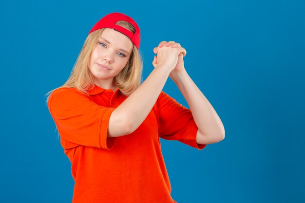 Entrega joven mujer vestida con polo naranja y gorra roja gesticulando con abrochado mirando confiado y orgulloso sobre fondo azul aislado