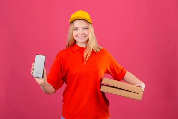 Entrega joven mujer vestida con polo rojo y gorra amarilla sosteniendo cajas de pizza mostrando teléfono móvil sonriendo amable sobre fondo rosa aislado