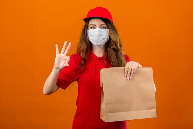 Entrega joven mujer vestida con polo rojo y gorra en máscara protectora médica de pie con el paquete de papel mostrando y apuntando hacia arriba con los dedos número tres mientras sonríe sobre naranja aislada