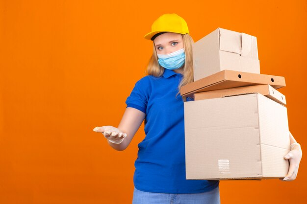 Entrega joven mujer vistiendo polo azul y gorra amarilla de pie con pila de cajas con la mano abierta esperando el pago sobre fondo amarillo oscuro aislado