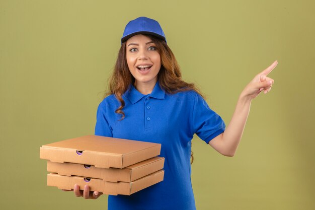 Entrega joven mujer vistiendo polo azul y gorra de pie con cajas de pizza mirando sorprendido sonriendo señalando con el dedo hacia el lado sobre fondo verde aislado