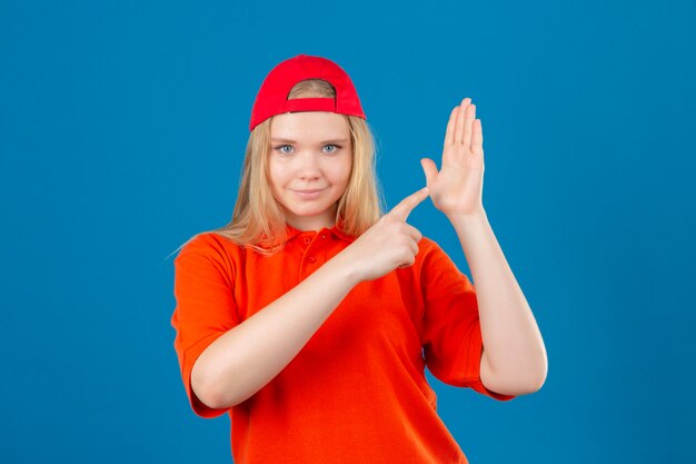 Entrega joven mujer vistiendo polo naranja y gorra roja apuntando a la mano abierta pal mirando a la cámara con una sonrisa en la cara sobre fondo azul aislado