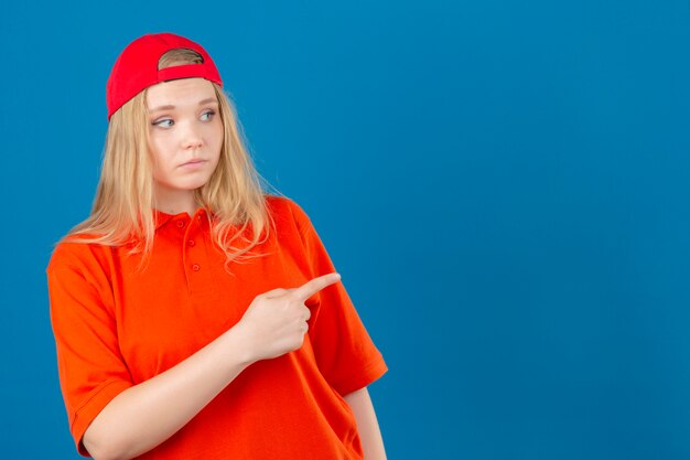 Entrega joven mujer vistiendo polo naranja y gorra roja mirando a un lado apuntando con el dedo índice a un espacio de copia sobre fondo azul aislado