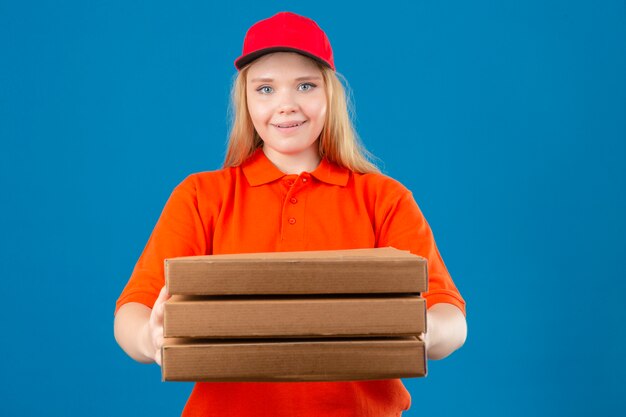 Entrega joven mujer vistiendo polo naranja y gorra roja sonriendo con cara feliz mirando a la cámara estirando una pila de cajas de pizza sobre fondo azul aislado