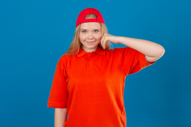 Entrega joven mujer vistiendo polo naranja y gorra roja sonriendo felizmente tocando la mejilla sobre fondo azul aislado